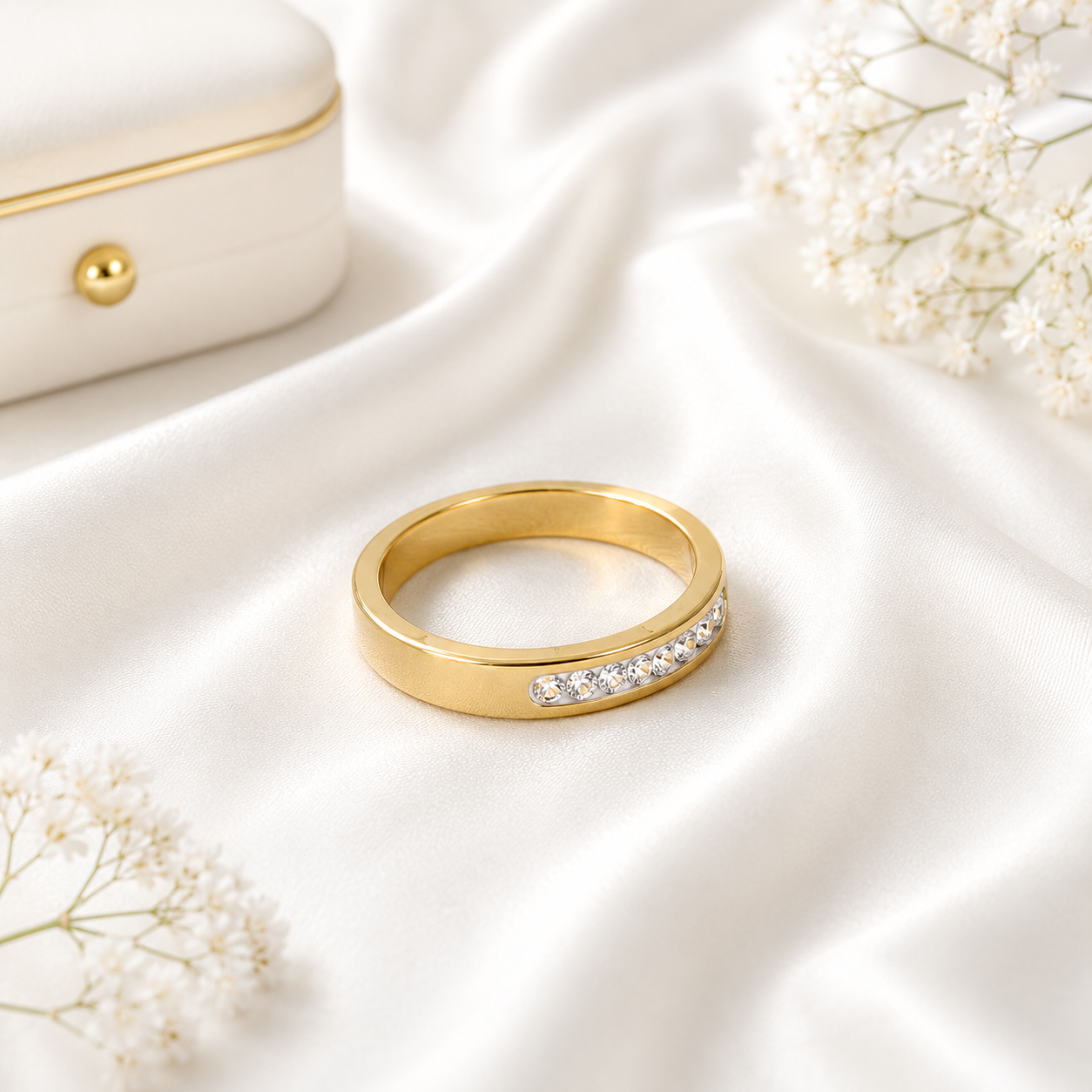Minimalist gold band ring with sparkling crystal accents displayed on soft white fabric beside a luxury jewelry box and delicate white flowers.