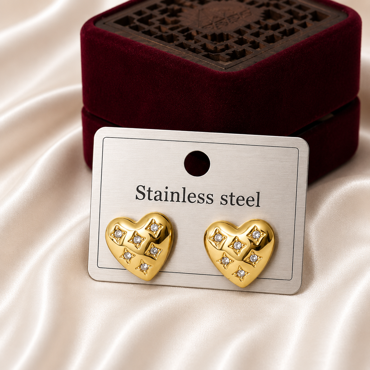 Gold-tone heart-shaped stud earrings with crystal accents displayed on a jewelry card against a luxury satin background with decorative gift box.