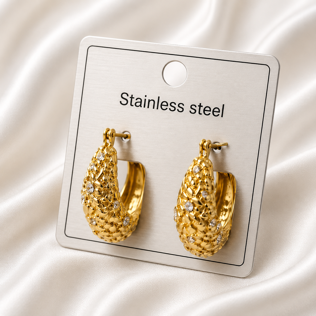 Gold-tone lattice hoop earrings with crystal accents displayed on a stainless steel jewelry card against a soft satin background.