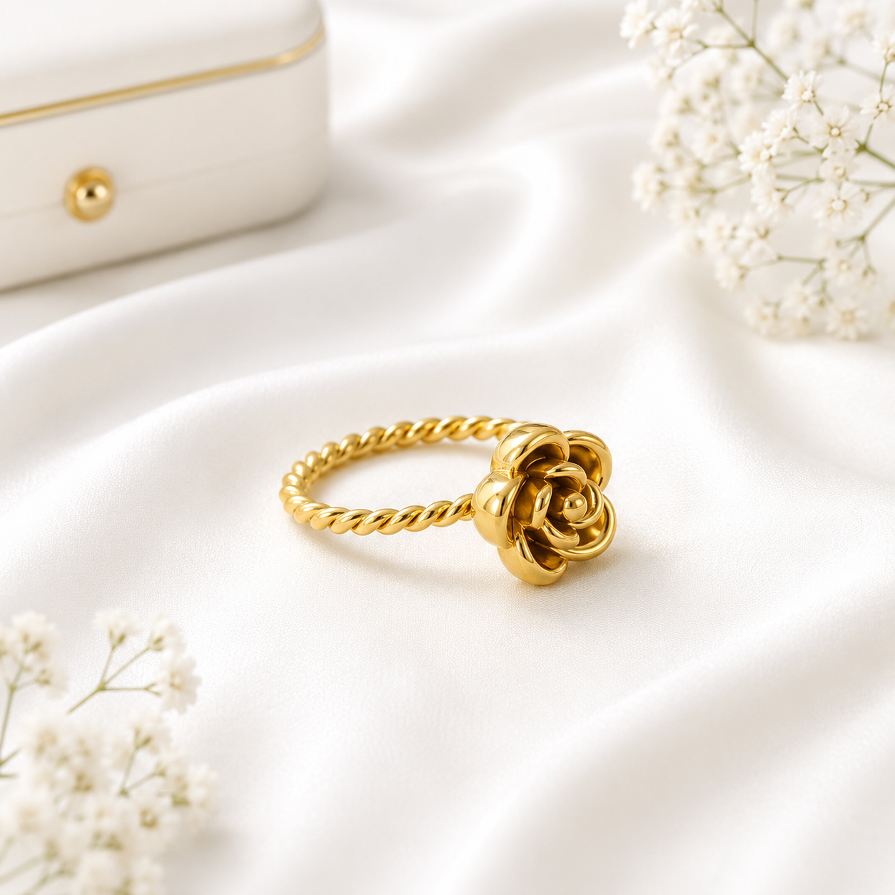 Gold rose flower ring with a twisted rope band displayed on soft white fabric with delicate white floral accents.